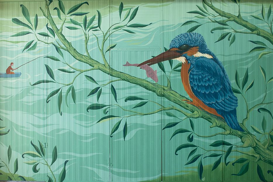 Dipinto murale - martin pescatore di colore blu e arancio appoggiato ad un ramo con un pesce rosa nel becco. Il ramo si affaccia su un laghetto e all'orizzonte c'è un pescatore su una barca azzurra